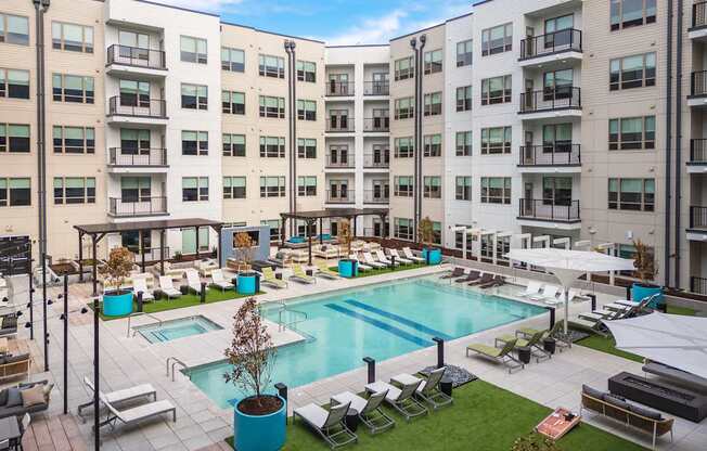 A large swimming pool surrounded by lounge chairs and umbrellas in front of apartment buildings.