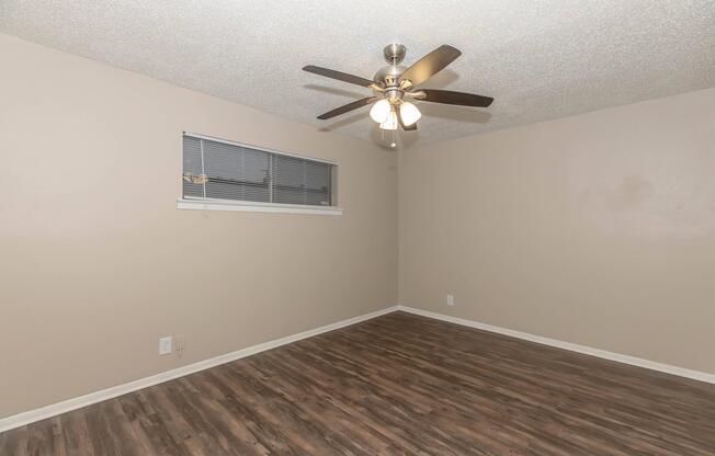 Empty room featuring a ceiling fan with four light fixtures, a window with blinds, and wooden flooring. The walls are painted a soft beige color, and there are no furnishings or decorations, creating a clean and minimalist aesthetic.