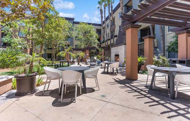 A patio with tables and chairs is surrounded by plants and trees at The Kitt at Warner Center Apartments, Woodland Hills 91303
