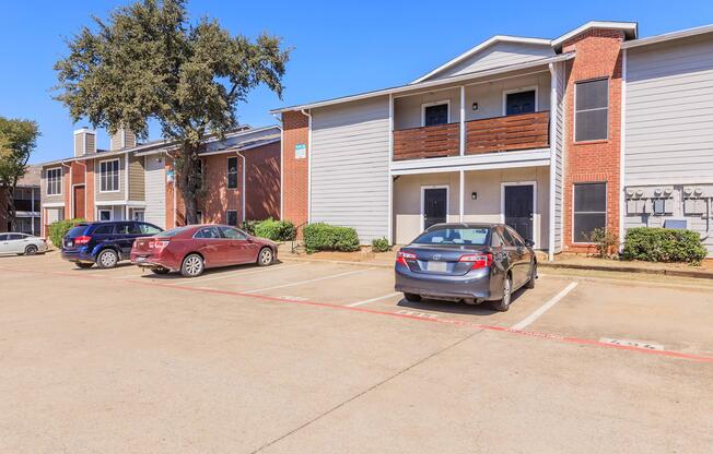 A view of a residential apartment complex featuring two buildings, parked cars in the foreground, and a clear blue sky. The buildings have a mix of brick and siding exteriors, with balconies visible on the upper levels. There are air conditioning units outside and landscaped areas around the parking lot.