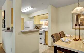 A welcoming kitchen and dining area here at Allegro featuring light wood cabinets, a breakfast bar with a raised counter, and a full appliance suite, opening to a dining space with a rectangular table, upholstered chairs, and a hanging pendant light, while neutral walls, soft carpeting, and warm finishes create a functional layout for everyday meals and entertaining.