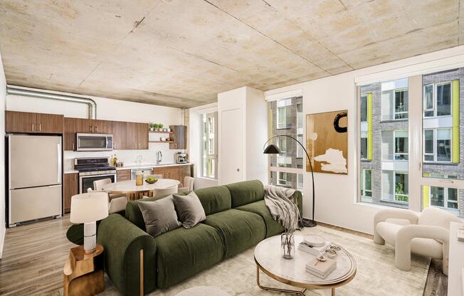 An open apartment home with concrete ceilings, expansive windows, and a streamlined kitchen, offering flexible living space filled with natural light.