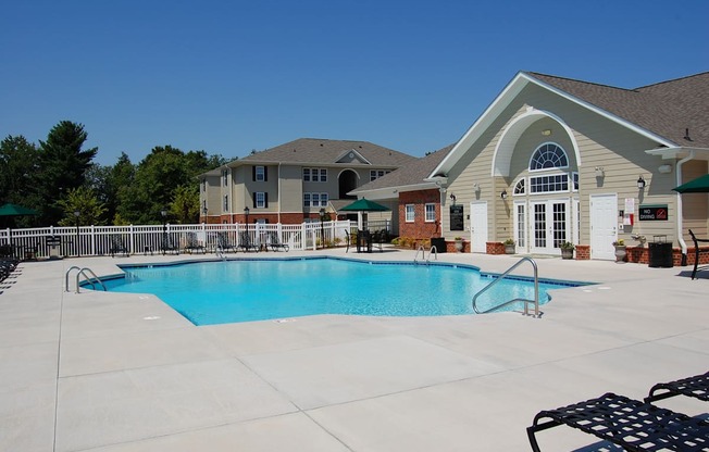 A large swimming pool in front of a building with a white fence and trees in the background.