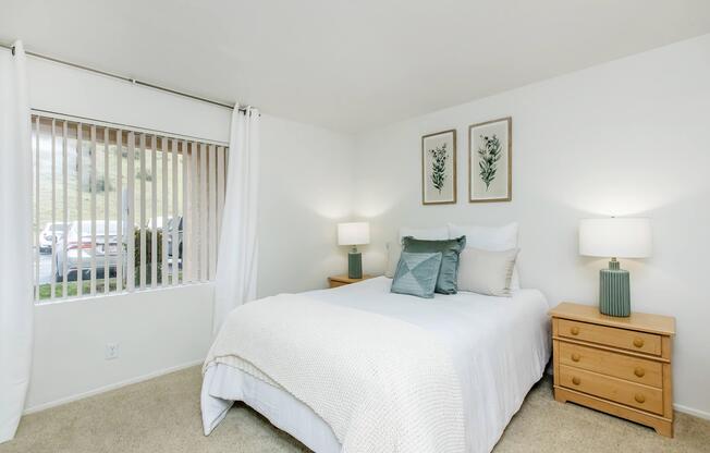 A cozy bedroom featuring a light-colored bedspread on a double bed, flanked by two wooden nightstands with lamps. Decorated with framed botanical prints on the wall and a window with beige blinds providing natural light. A neutral carpet enhances the inviting atmosphere.