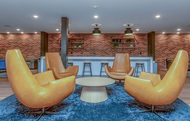 A modern lounge area featuring four stylish orange armchairs arranged around a round wooden coffee table, situated on a soft blue rug. In the background, there is a bar-like counter with high stools against a textured brick wall, creating a cozy and inviting atmosphere.