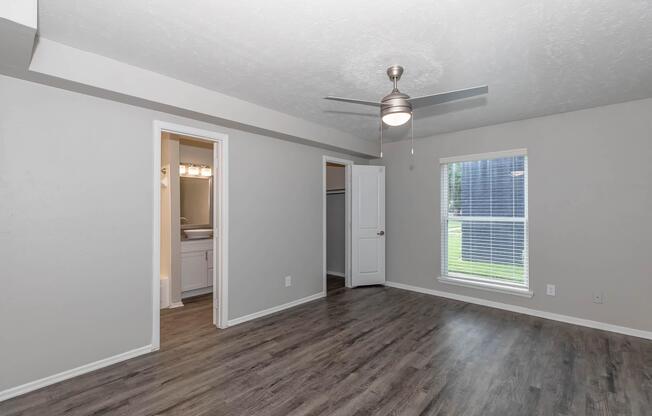 A spacious room with gray walls and a ceiling fan. It features a window with natural light, a closet with sliding doors, and an entryway leading to a bathroom. The flooring is wood-like laminate, creating a modern and inviting atmosphere.