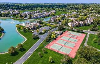A tennis court is located in a grassy area near a lake and houses.