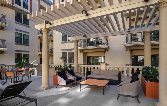 A cozy outdoor lounge area featuring a pergola, comfortable seating, and a coffee table. Surrounded by potted plants, it is located within a well-designed building with balconies. Sunlight filters through the slats of the pergola, creating a welcoming atmosphere for relaxation or socializing.
