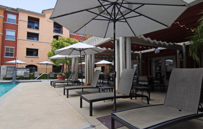 A patio with chairs and umbrellas is surrounded by a pool and apartment buildings.