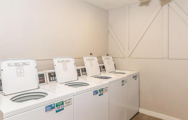 A clean laundry room featuring four white top-loading washing machines, each equipped with control panels. The room has light-colored walls and a ceiling fan, creating a bright and spacious environment.