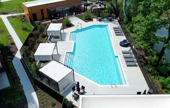 A sparkling resort-style pool near trees with lounge chairs, tables, chairs, and 3 shaded lounge areas.
