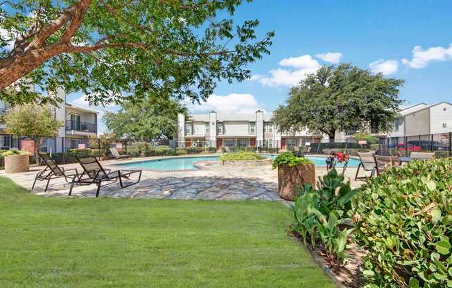 A resort-style outdoor pool area here at High Plains with a sparkling swimming pool, lounge chairs, and a stone pool deck surrounded by lush green landscaping. Mature trees provide shade, and nearby apartment buildings create a calm, inviting community atmosphere.