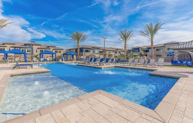 A bright outdoor swimming pool area featuring a large blue pool with lounge chairs and cabanas. Palm trees line the space, and modern buildings are visible in the background under a blue sky with wispy clouds. The pool includes a shallow section and well-maintained tiled edges.