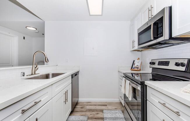 Modern kitchen featuring white cabinetry, stainless steel appliances including an electric stove and microwave, a stainless steel sink, and light-colored countertops. The floor has a wood-like finish, and there are decorative mats in front of the sink and stove. The space is bright and airy with neutral tones.
