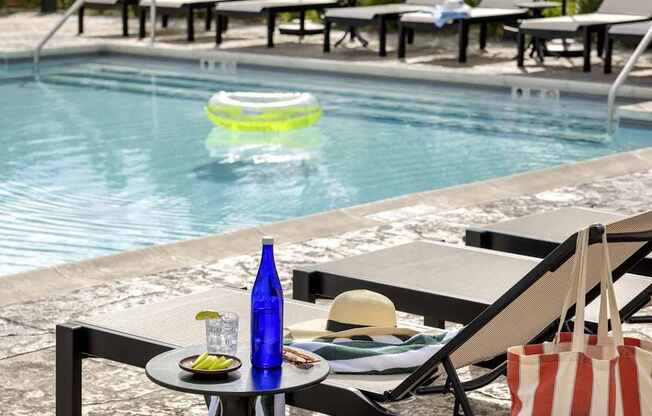 A poolside table with a hat, a bottle, a plate of food, and a bag.