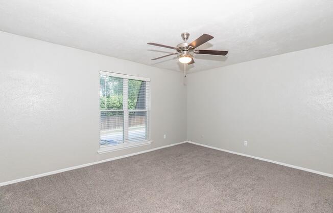 A spacious, empty room with light gray walls and a ceiling fan. The floor is covered with beige carpet, and there is a window letting in natural light. The window has a view of a green space outside, and the overall ambiance is bright and open.