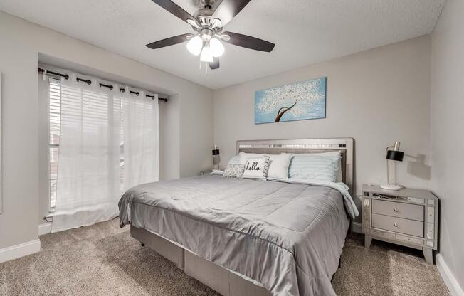 A cozy bedroom featuring a gray bedspread and decorative pillows, a large window with sheer curtains letting in natural light, and a modern ceiling fan. A piece of artwork hangs above the bed, and a small nightstand with a lamp is positioned beside it. The room has soft carpeting and light gray walls.