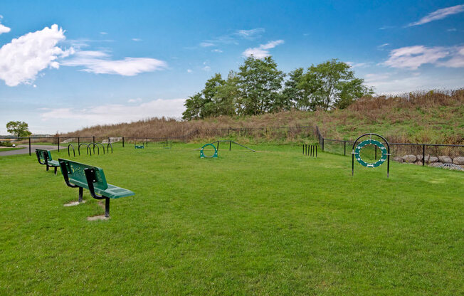 A dog park here at The Park featuring green grass, agility obstacles, and benches, creating a spacious, pet-friendly area with playful features and a relaxing place for residents and their dogs.