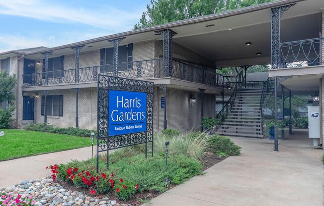 A view of Harris Gardens, a residential building showcasing a welcoming sign with the text "Harris Gardens - Unique Urban Living." The structure features multiple levels with decorative railings, landscaped greenery, and flower beds in front, creating a pleasant community atmosphere.
