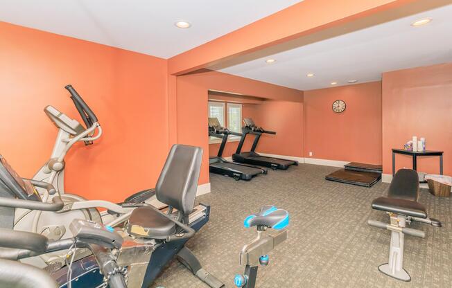 A well-lit home gym featuring exercise equipment including treadmills, an elliptical machine, and a weight bench. The walls are painted orange, and there is a mirror on one side reflecting the workout area. A clock is mounted on the wall, and a small table holds fitness supplies.