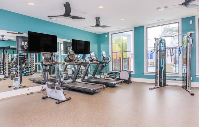 A modern gym interior featuring three treadmills, a stationary bike, and a multi-gym weight machine. Large mirrors and two wall-mounted televisions enhance the space, which has bright turquoise walls and ample natural light from windows. The flooring is a durable, textured material suitable for exercise.