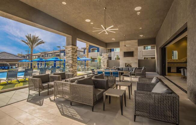 An outdoor lounge area featuring comfortable seating with modern, wicker-style furniture. The space is covered, with large windows offering a view of a swimming pool and surrounding palm trees. Blue umbrellas provide shade, and there's a large flat-screen TV mounted on the wall.