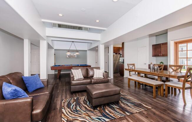 A spacious living room featuring a brown leather sofa with blue cushions, a patterned area rug, and a coffee table. An adjacent dining area has a wood table with chairs, and a kitchen area visible in the background. Natural light fills the space, highlighting modern decor and an inviting atmosphere.