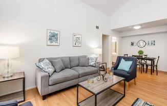 A cozy living room featuring a gray sofa with decorative pillows, two accent chairs, a glass coffee table, and light wooden flooring. The walls are adorned with framed art pieces, and there is a dining area visible in the background, showcasing a round table with chairs. Soft lighting creates a warm atmosphere.