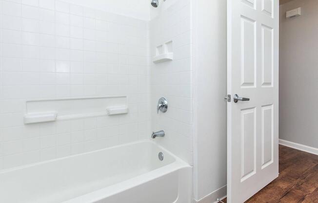 A clean, modern bathroom featuring a white bathtub, tiled wall with built-in shelves, a chrome faucet, and a showerhead. The door to the bathroom is partially open, revealing a light gray wall and wood flooring.
