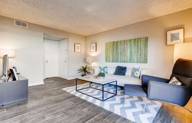 A living room with a black and white chevron rug, a black leather chair, a white couch with blue and white pillows, a wooden coffee table, and a white wall with two framed pictures.