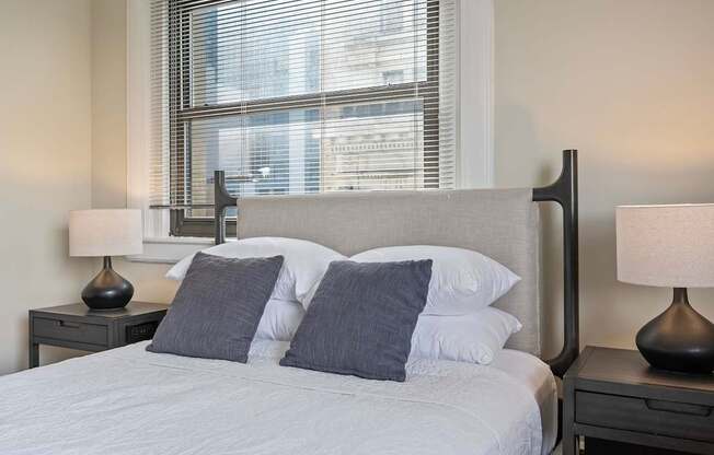 A bed with white sheets and grey pillows is in front of a window with blinds.