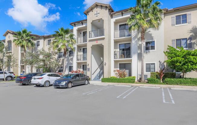 A multi-story residential building featuring a light-colored facade, balconies, and tropical landscaping. Several parked cars are visible in the foreground, with palm trees and shrubs enhancing the exterior. The sky is partly cloudy, adding to the bright atmosphere of the setting.