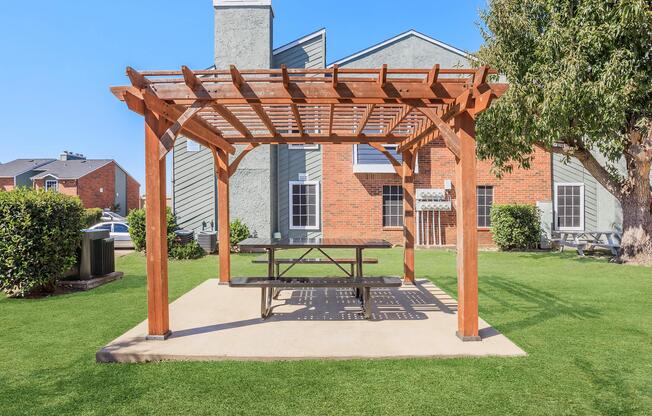 A wooden pergola with a picnic table underneath, situated on a grassy area. In the background, there are two residential buildings with brick and gray exteriors. The sky is clear and blue, creating a bright and inviting atmosphere.