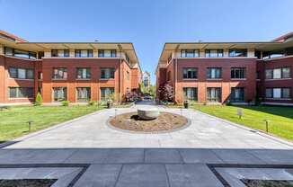 A courtyard with a fountain in the middle of a paved area with two buildings in the background.