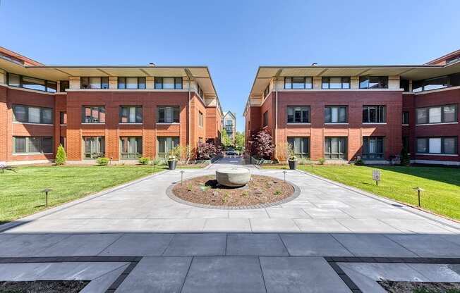 A courtyard with a fountain in the middle of a paved area with two buildings in the background.