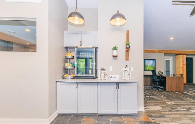 A modern interior space featuring a light gray cabinet with a countertop, a glass display stand filled with snacks, and decorative items like potted plants and lanterns. Pendant lights hang above, and soft flooring leads to a connected workspace with wooden accents.