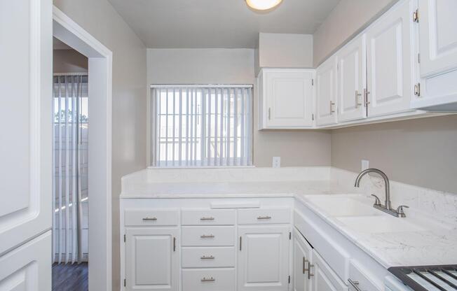 Bright and modern kitchen with white cabinetry, a marble countertop, and a stainless-steel sink. The window above the sink features vertical blinds, allowing natural light to enter. A glimpse of an adjoining room is visible through an open doorway. The flooring is dark wood-like laminate.