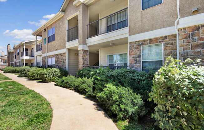A well-maintained apartment building exterior here at Mission Green showcasing stone and stucco architecture, private balconies, large windows, neatly trimmed landscaping, and a curved concrete walkway bordered by green grass and shrubs beneath a bright blue sky.