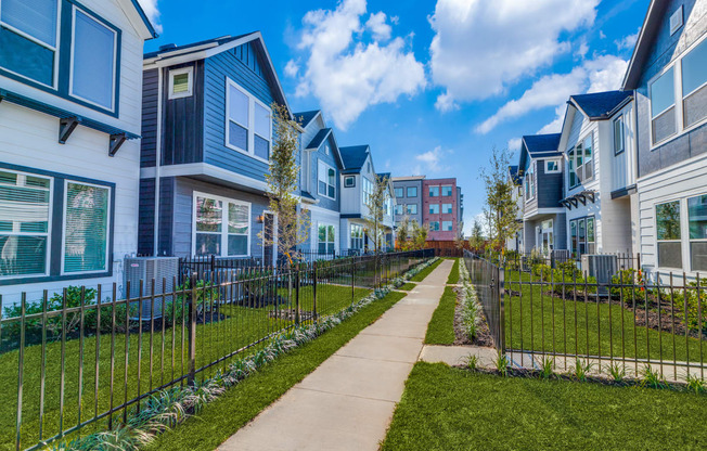 Houses for Rent in Houston, TX - East Heights - Exterior View of Apartment Homes with Fences, Manicured Grass Yards, Concrete Walking Path, and Trees.