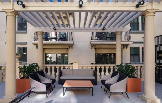 Outdoor lounge area with a modern design featuring a pergola overhead supported by columns. There are four comfortable chairs arranged around a coffee table, with potted plants nearby. The background shows a building with balconies and windows, creating a cozy and inviting atmosphere.
