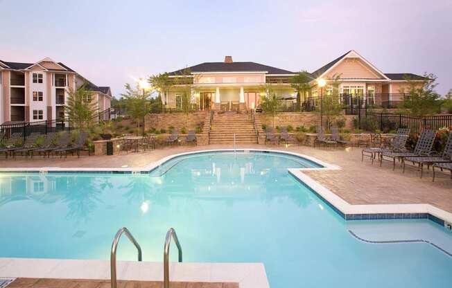 A swimming pool in front of a house with a patio and chairs.