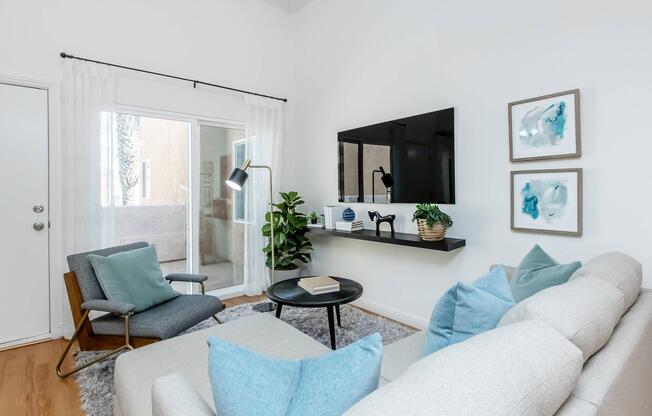A modern living room with light-colored walls and hardwood flooring. It features a comfortable gray sofa with blue cushions, a black round coffee table, a stylish armchair, and a potted plant. A flat-screen TV is mounted on the wall above a floating shelf with decor, and there are glass doors leading to an outdoor area.