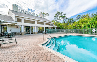 Swimming pool with large tanning deck, table with umbrella, lounge chairs, clubhouse in background surrounded by native landscaping