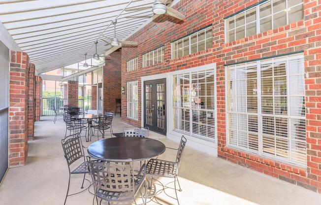 An outdoor seating area featuring black metal tables and chairs under a covered patio with brick walls and large windows. The space is bright and airy, designed for relaxation or gatherings.