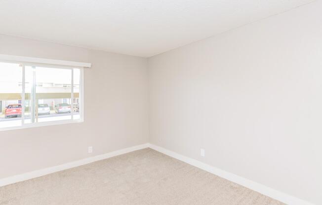 A light and airy empty room with beige carpet and pale gray walls. A window with a white blind allows natural light to flow in, showcasing a view of parked cars outside. The room has a clean, minimalist aesthetic, suitable for various uses or decorating styles.