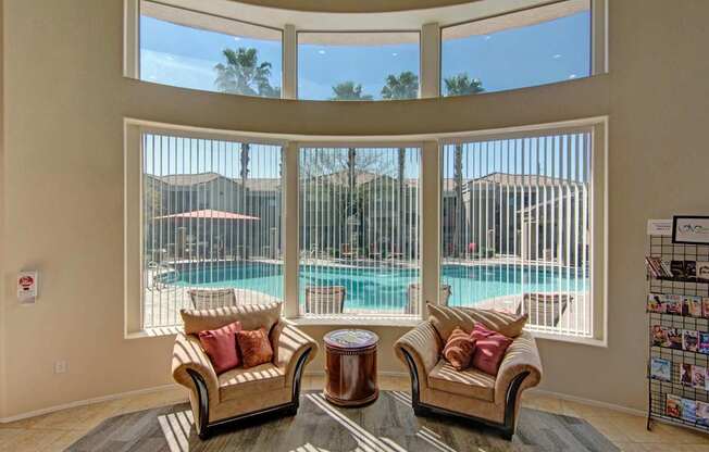 A welcoming lounge space here at Dobson Towne Center featuring two plush accent chairs with warm-toned pillows arranged beside a round wood side table, set against expansive curved windows with vertical blinds that frame views of the sparkling swimming pool, palm trees, and surrounding buildings, all illuminated by abundant natural light for an inviting, relaxed atmosphere.