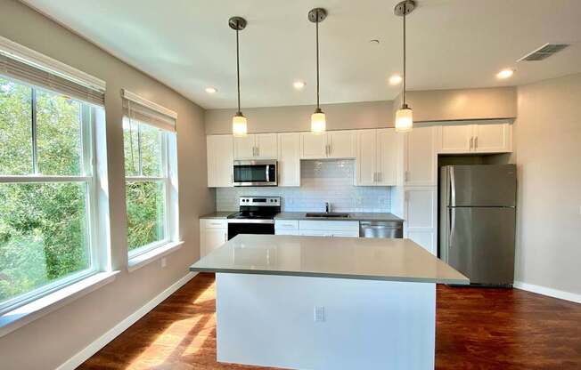 A well-lit open concept kitchen with two large windows, recessed and pendant lighting, hardwood-style flooring, white cabinets, subway tile backsplash, and stainless steel appliances.