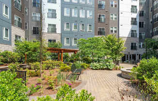 The Residences at Crosspoint central courtyard at 42 Wellman Street in Lowell, MA features paved walkways, raised garden beds, green shrubs, benches, and surrounding multi-story buildings clad in stone and metal siding.