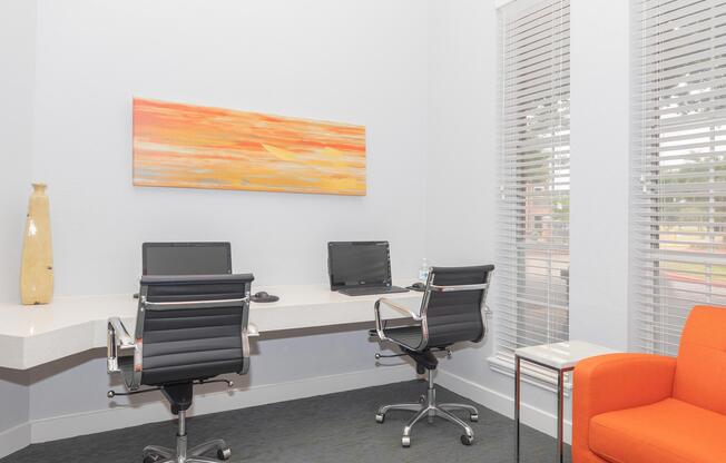 A modern office space featuring two sleek black chairs at a white desk with two computers. There is an orange armchair in the corner and a colorful abstract painting on the wall. The room has large windows with blinds letting in natural light, creating a bright and inviting atmosphere.