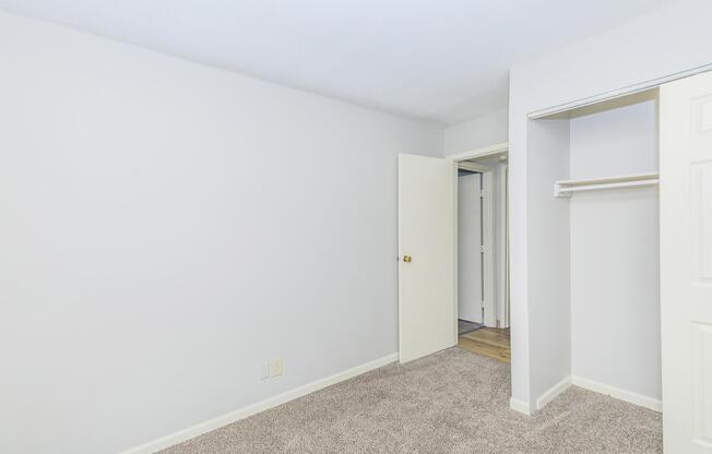 A clean, empty bedroom with light gray walls and plush carpet. A white sliding closet door is partially open, revealing shelving. An open door leads to a small hallway, indicating access to another area. The overall atmosphere is bright and spacious, suitable for personalization.